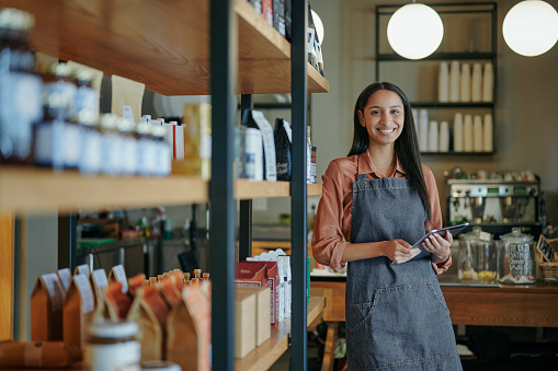 Portrait of a smiling young female deli owner standing with a digital tablet by shelves of artisanal merchandise in his store