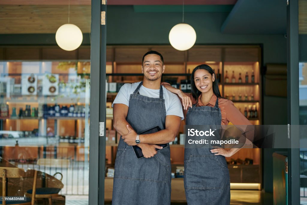 Smiling young deli owners standing at their store entrance Portrait of two smiling young delicatessen owners standing together at the front entrance to their store Small Business Stock Photo
