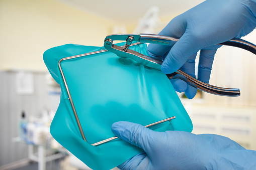 Dentist's hands in latex gloves with a dental hole punch, a rubber dam and a metal frame with a dental clinic on the background. Medical tools concept.