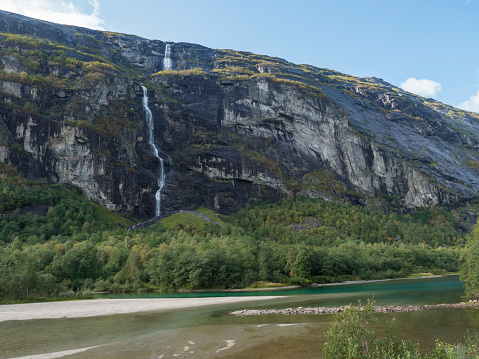 Tall waterfall with turquoise Rauma river at Romsdalen valley with rocks and green forest. Blue sky white clouds background. Norway summer landscape.