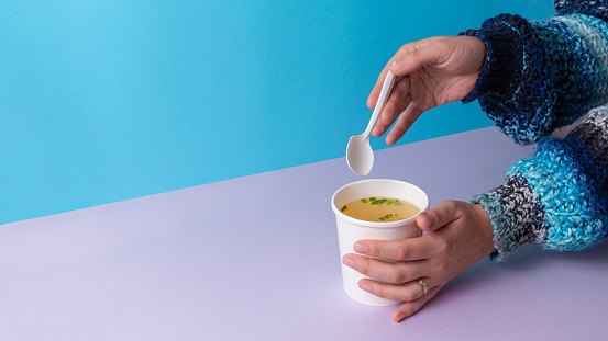 Famele eating a Chicken broth soup in takeaway cup with spoon. Minimalistic Blue and purple background with copy space