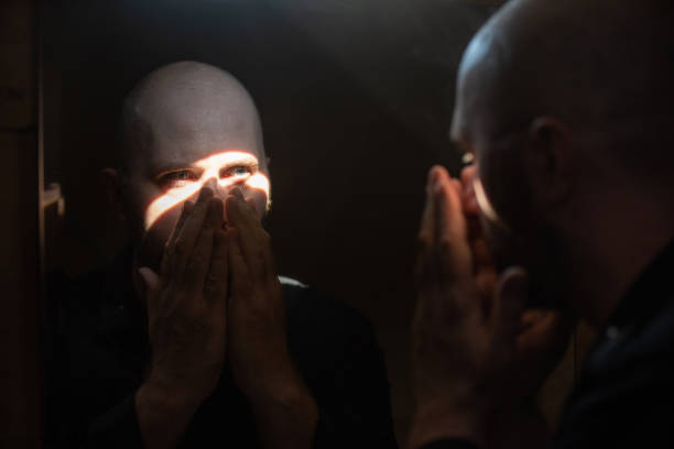 A man in a dark bathroom, closing his face with his hands, we see his reflection in a mirror in front of him in a sunbeam light, probably coming through the gap in a wall. A scene of religion, a man prays alone.