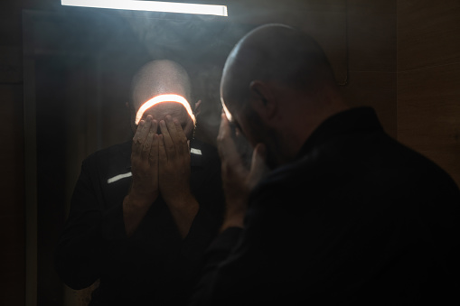A man closes his face with hand, standing in front of the mirror in a dark room. A scene of religion, a man prays alone.