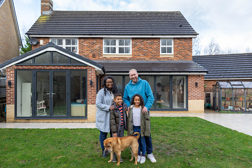 Multi-racial family standing proudly in front of the home in the garden. They are all looking at the camera with positive emotion. The dog is walking around their legs.