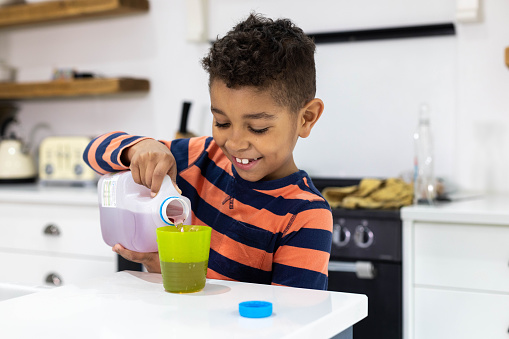 A young boy pouring juice out of a plastic carton into a green cup. He is looking down concentrating on what he is doing.