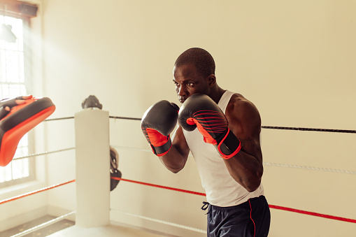 Photo of a boxer training with a punching bag in a boxing hall. There is a boxing ring behind him.