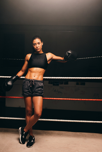 Fit young woman looking at the camera while standing against the ropes in a boxing ring. Female boxer training in a fitness gym.