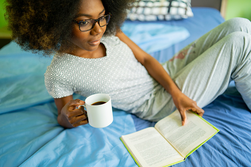 Close up of a young woman lying back on a bed, holding a book in one hand and a black coffee in a mug in the other, wearing eyeglasses and pajamas as she relaxes