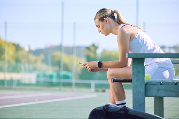 Fit tennis player with phone checking fitness goal progress on a sports exercise app online while taking a break at the court. A sportswoman checking messages on cellphone and waiting for a coach Fit sports woman with phone checking fitness goal progress on a modern exercise app online while taking a break at the tennis court. A sportswoman checking messages on cellphone and waiting for coach tennis coach stock pictures, royalty-free photos & images