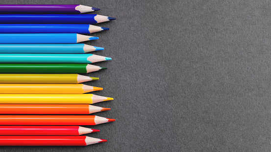 Horizontal row of colorful pencils on grey paper background. School supplies in colores of rainbow on blackboard. Kid's stationery with black copy space. Back to school backdrop.