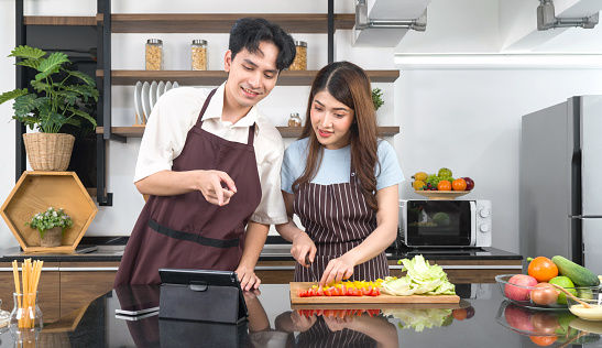 Young asian woman dressed in an apron holding knife slices the vegetable into pieces on a wooden chopping board. Her boyfriend pointing at cooking recipe on tablet computer. Atmosphere in a kitchen.