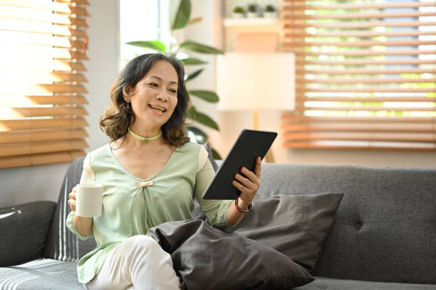 istock Middle aged woman in casual clothes drinking hot coffee and using digital tablet on couch at home. 1416897877