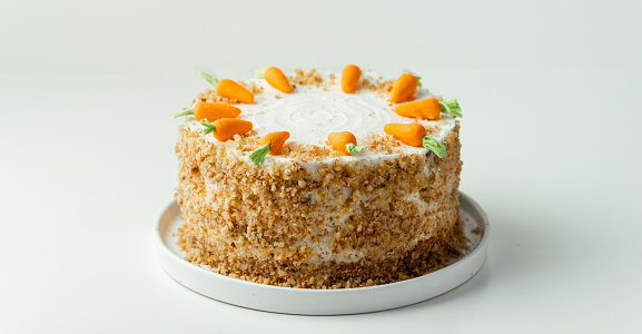 Delicious carrot cake decorated with mastic sweet carrots. Homemade carrot cake with yellow crumbs in the white plate on the white background
