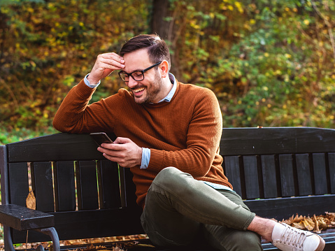 Smiling man sitting at park bench and using smartphone