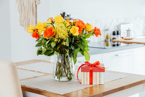 Yellow and orange roses in a vase and gift box with red ribbon on kitchen counter table with modern kitchen background. Gift for Thanksgiving day, birthday, Mother's or Valentine's day. Copy space.