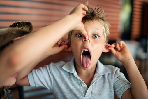 Portrait of funny little boy making face at the camera. Sister is helping him with the nose.\nNikon D800