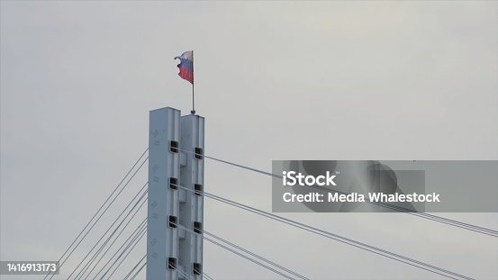 istock Russian Federation flag fluttering in the wind on the top of the bridge. Stock. Russian flag on the top of a bridge on grey, cloudy sky background. 1416913173