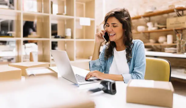 Happy entrepreneur speaking on the phone while working on a laptop in her warehouse. Online store owner making plans for product shipping. Creative businesswoman running an e-commerce small business.