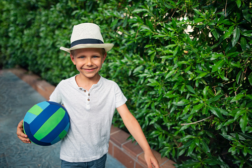 Little boy is going to play soccer/football in the park. The boy aged 7 is holding the soccer ball and smiling at the camera. \nNikon D800