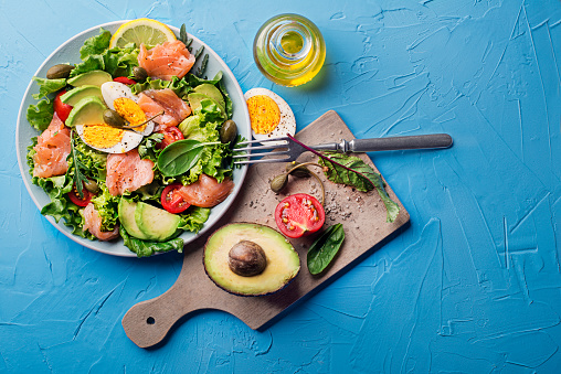 Fresh green lettuce salad with smoked salmon, cherry tomatoes, egg and avocado on blue table background
