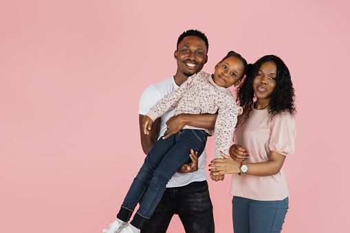 Cheerful african man and his wife holding excited daughter on hands. Woman and her husband looking and posing at camera, standing isolated over pink studio background, panorama, banner free copy space