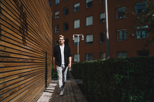 Confident male in glasses dressed in grey pants black jacket and white t shirt walking along wooden wall in city at daytime