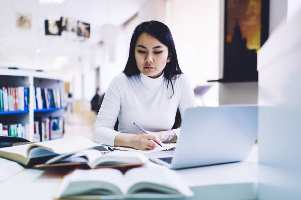 istock Asian woman reading book while sitting at desk and taking notes in notebook in library 1416953121