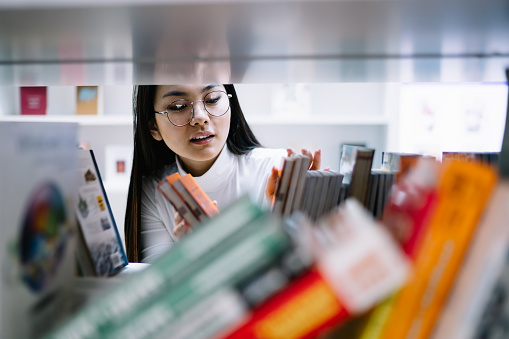Black haired young Asian female in white top and glasses selecting book on shelf of bookcase in library