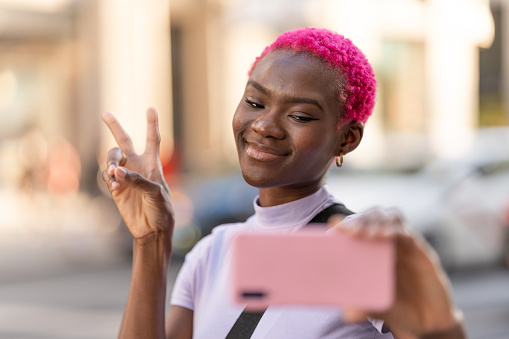 Smiling young stylish afro woman taking selfie while gesturing victory in the street