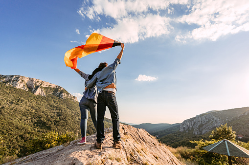 Beautiful young happy couple waving a German flag at the top of the mountain.