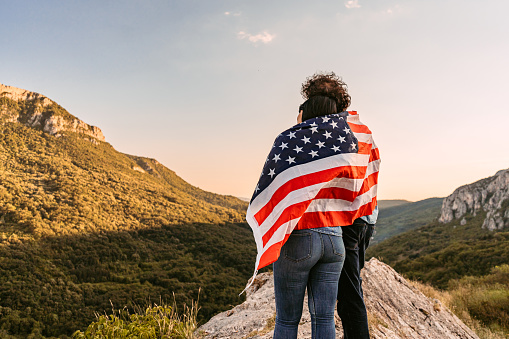 Beautiful young happy couple wrapped in an American flag at the top of the mountain.