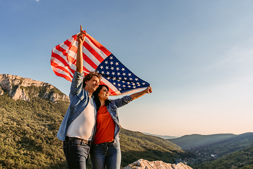Beautiful young happy couple waving an American flag at the top of the mountain.