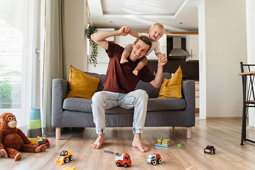 Happy father carrying his laughing little blond son on neck and sitting on couch in living-room surrounded with toys, spending time together on weekend. Young dad playing with his toddler boy