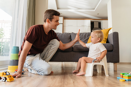 Potty training. Young father assisting his little baby boy using pot in living room. Cute baby giving high five to his dad sitting on potty at home. Dad teaching his son how to use potty
