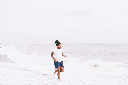 Full length African American male in sportswear jogging on sandy coast near waving sea during summer stroll in sunny day