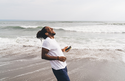 From above side view of laughing African American male in casual clothes smiling and listening to music on mobile phone