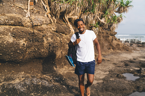 Happy ethnic guy in casual summer clothes smiling with backpack on shoulder while standing on stony sea coast in summer day