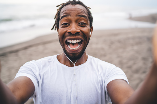 Happy African American male runner in sportswear with headphones smiling and looking at camera while resting on sandy beach after training
