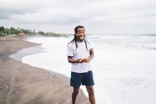 Excited laughing bearded black guy with dreadlocks in casual clothing enjoying walk on sandy beach near ocean waves on cloudy day