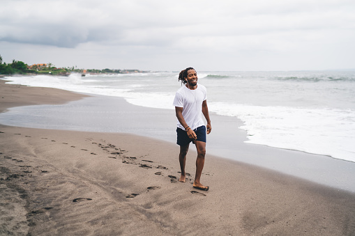 Full length ethnic male in white shirt and blue shorts with earphones walking barefoot on coastline while ocean waves washing sand