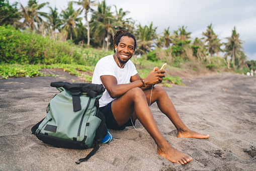 Happy African American male in casual clothes sitting on sandy seashore and browsing web while spending time in tropical resort