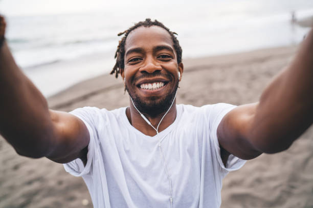 Joyful black man holding camera and taking selfie on beach