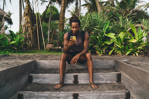 Full body of young African American male in casual clothes sitting on stair in tropical country and browsing mobile phone in soft daylight