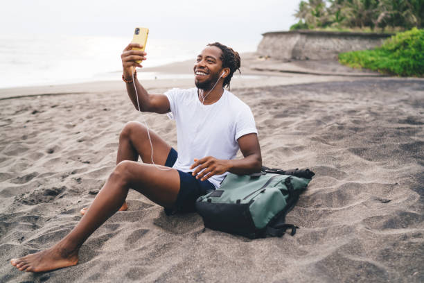 homem negro alegre tendo chamada de vídeo na praia - internet computer monitor discussion surfing the net - fotografias e filmes do acervo