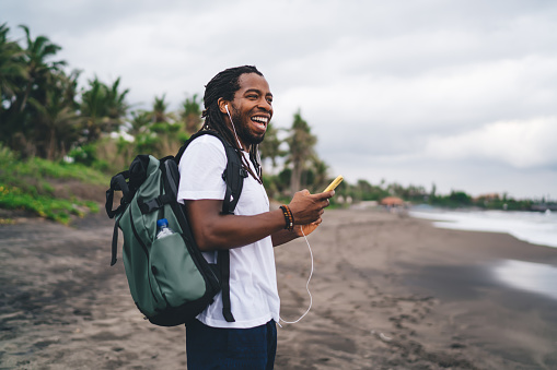 Side view joyful African American male in casual shirt using mobile phone and earphones while standing with backpack on exotic beach and looking away happily