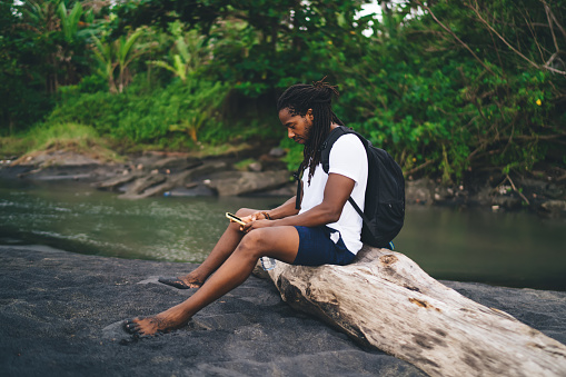 Side view full body concentrated African American male hiker in summer wear sitting on dry tree trunk near shallow river in forest and browsing mobile phone