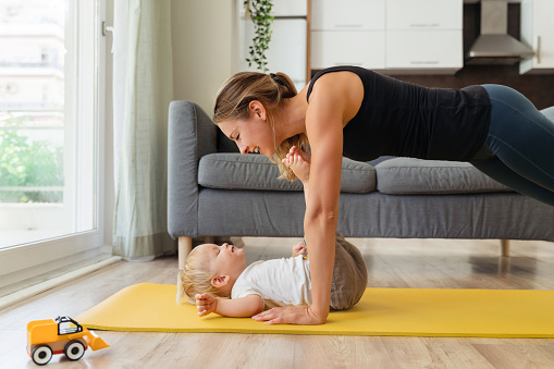 Side view image of mother practicing yoga standing in plank position above his cute baby boy lying on mat, reaching his legs to her face. Mom working out with her toddler. Happy healthy motherhood