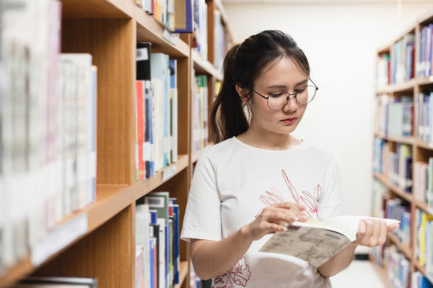 Adult education concept and leisure activity, Asian woman reading a book, between bookshelves in the library. Adult education concept and leisure activity, Asian woman reading a book, between bookshelves in the library. 5. Library Assistant stock pictures, royalty-free photos & images
