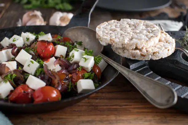 Delicious warm fitness salad with roasted cherry tomatoes, spanish onion, herbs, olive oil and sheep cheese. Served with rice cakes on wooden table