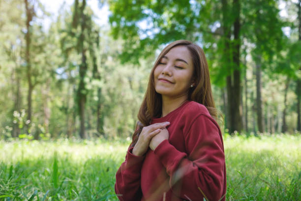 portrait d’une jeune femme les yeux fermés posant les mains sur sa poitrine dans le parc - spirituality hand on heart meditating women photos et images de collection